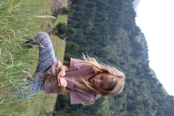 A young child with blonde hair, wearing a pink shirt and patterned leggings, is gently observing blades of grass on a lush green mountain slope